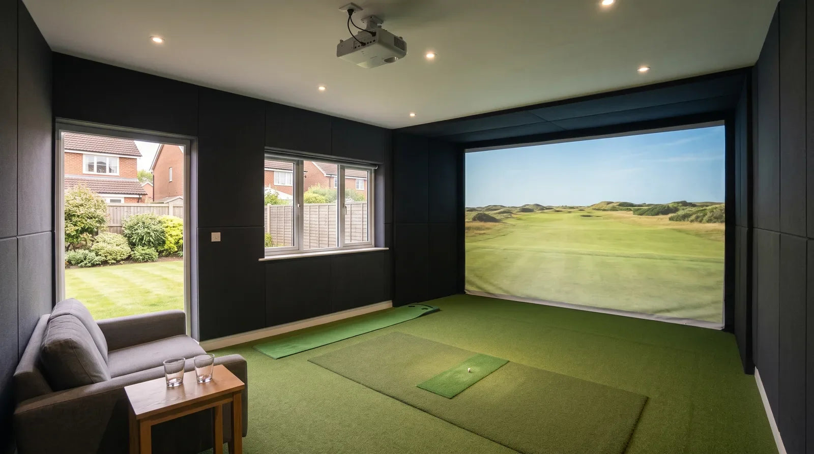 A finished golf simulator room in a British house, with dark acoustic panels on the walls, an impact screen showing a links fairway, a hitting mat on turf, a putting strip, and an armchair and side table at the back of the room